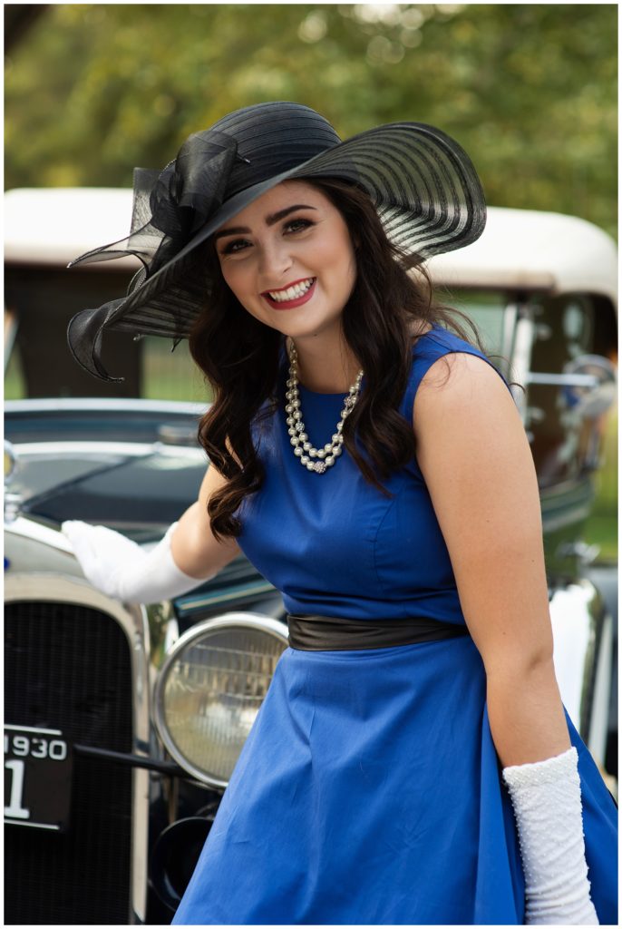 girl in blue dress and black hat laughing in front of vintage car