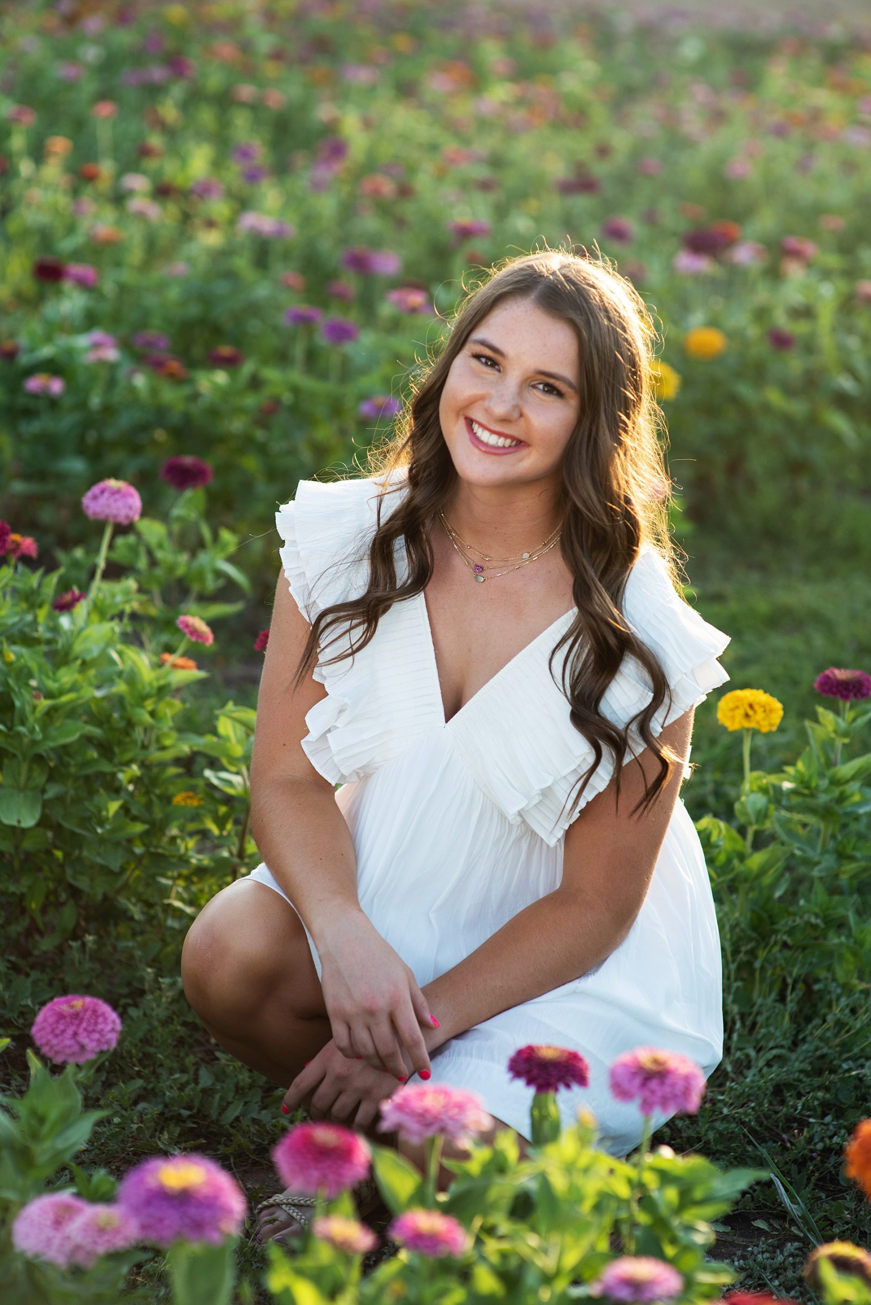 high school senior girl in white dress brown hair sitting in field of zinnias senior pictures