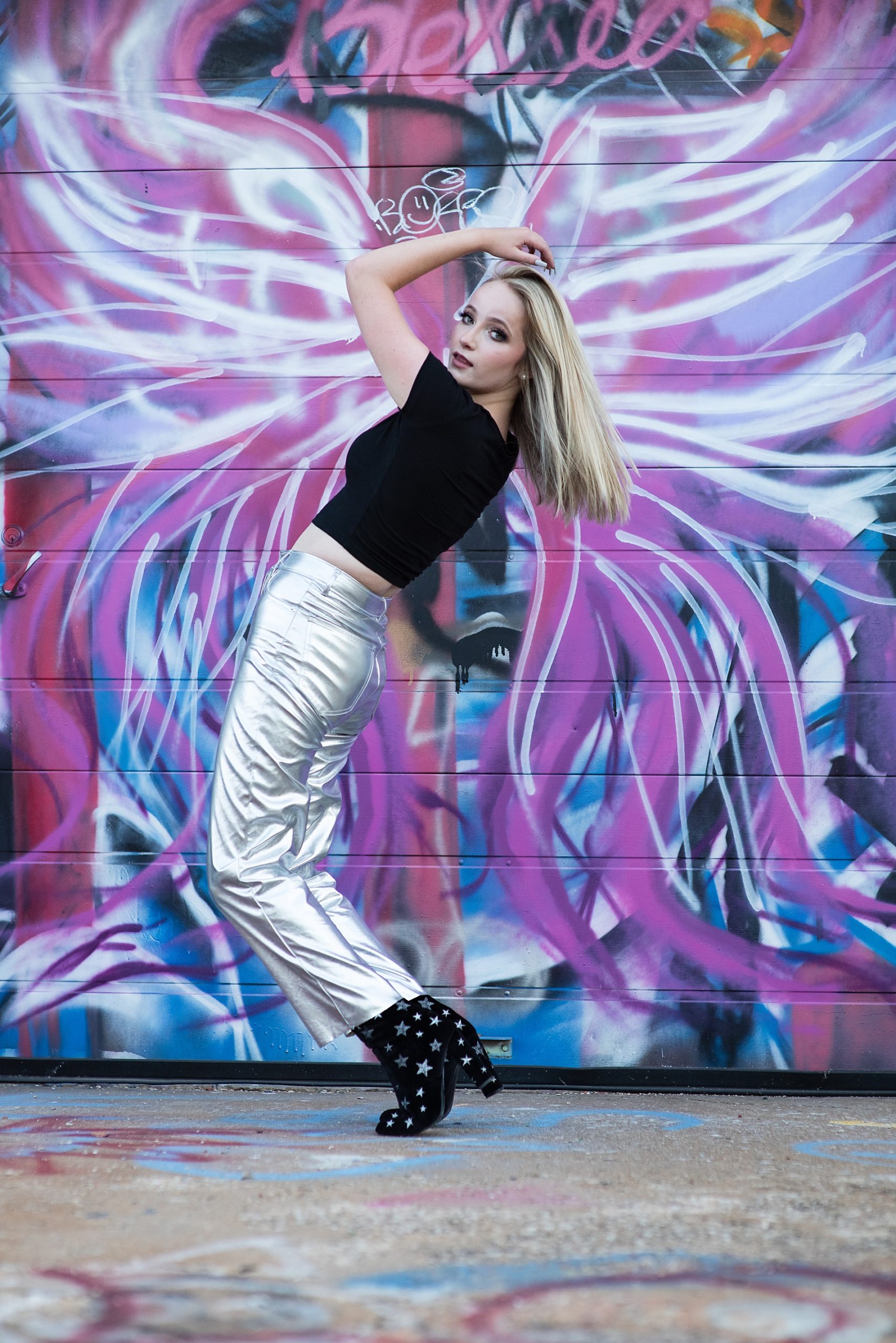high school senior girl in silver pants, black shirt, black boots in front of graffitti wall senior pictures