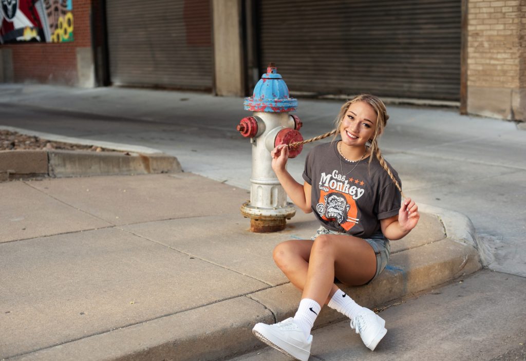 high school senior in jean shorts, graphic tee sitting in front of fire hydrant senior pictues