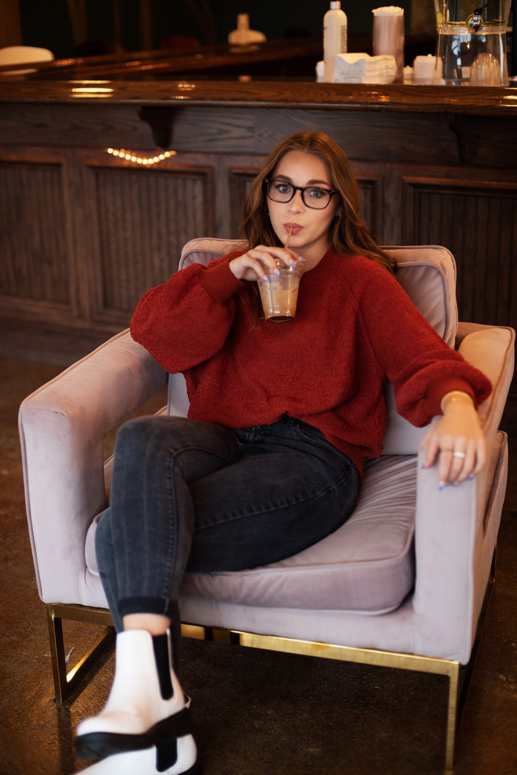 high school senior girl in black jeans, white boots, maroon top, sitting in pink chair sipping coffee senior pictures