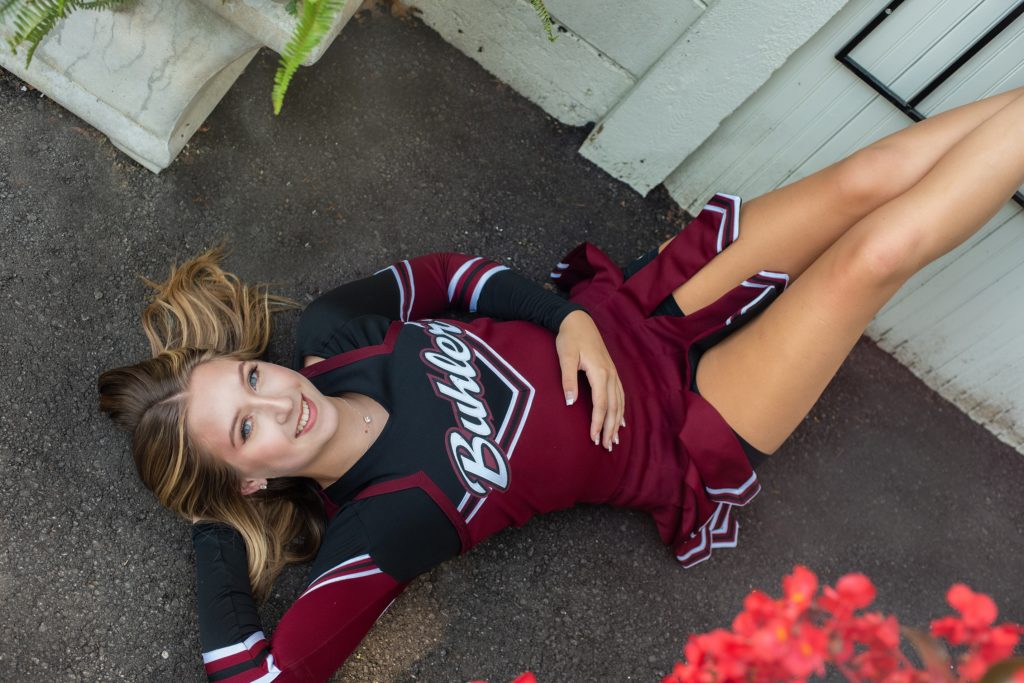 high school senior in cheer uniform lying down next to building senior pictures 