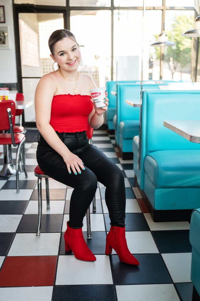 high school senior girl in red top and black pants, red boots in a diner photoshoot